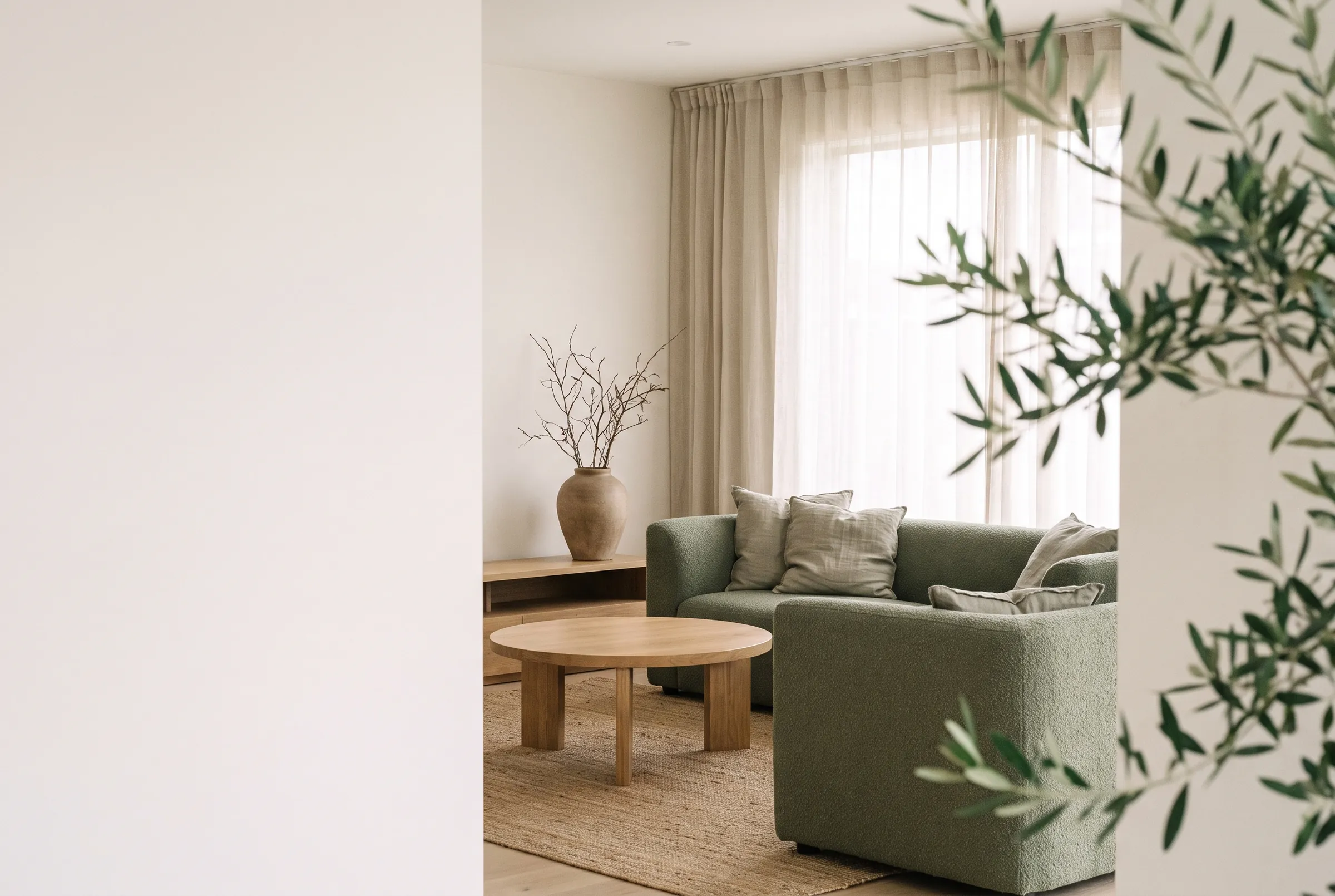 A calm living room with a sage sofa, light oak table and an olive tree by the window